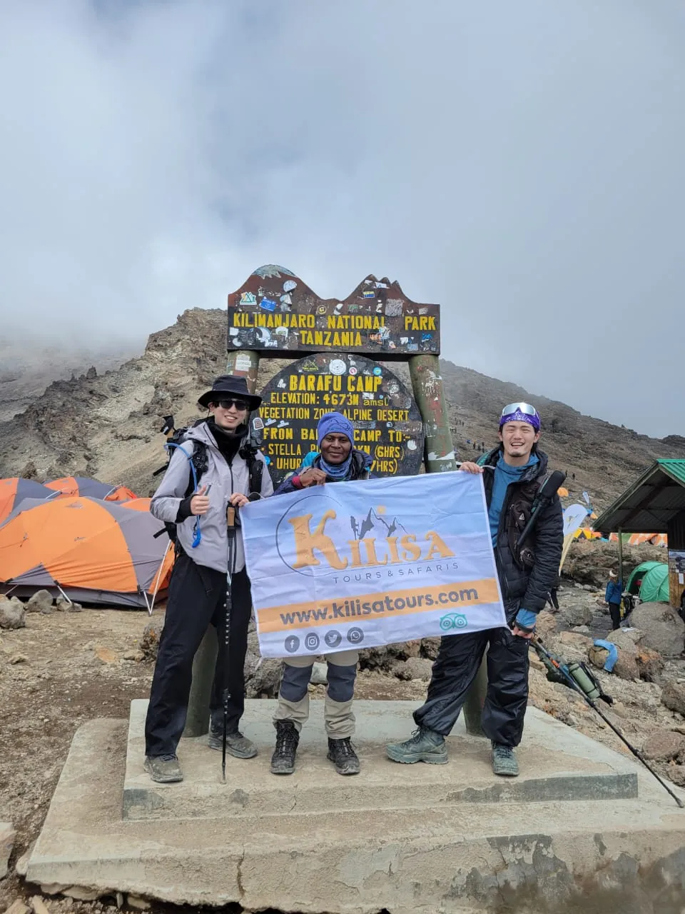 Three climbers holding a Kilisa Tours banner at the Barafu Camp sign, base camp for the Kilimanjaro summit