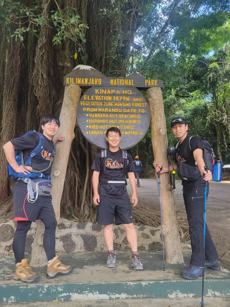 Three trekkers at the Marangu Gate sign, 1,879 m, at the start of the Marangu route in Kilimanjaro National Park